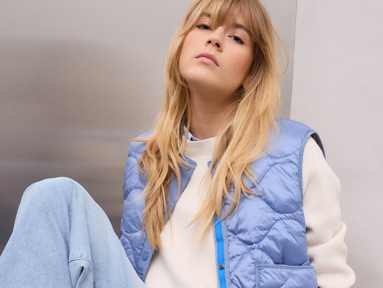 The woman is wearing a white jumper with a blue blouse underneath, a light blue waistcoat and jeans.