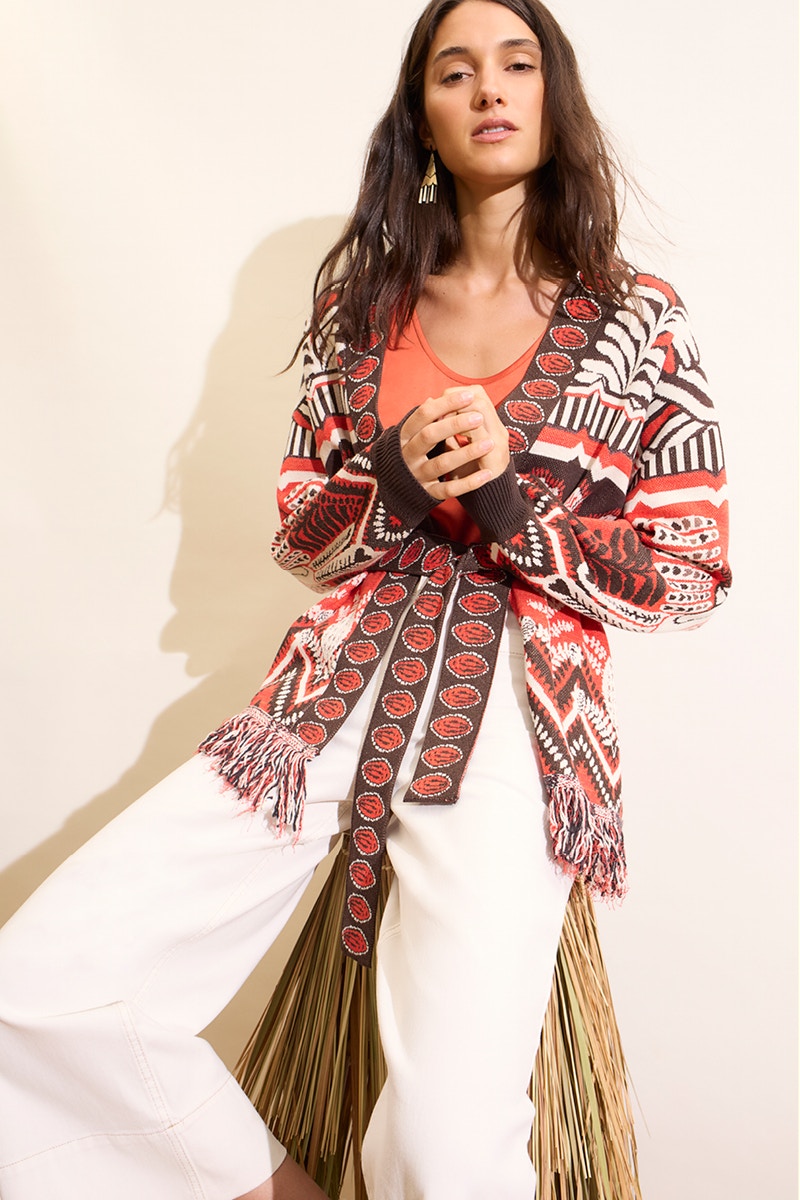 Woman wearing a patterned cardigan in brown, orange and white, an orange shirt and white trousers.