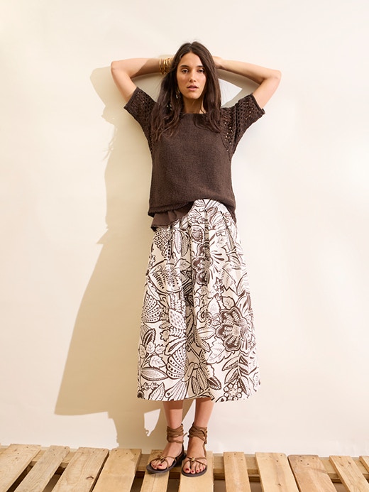 Frau mit braunem kurzarm Pullover und gemusterten weißen Rock mit braunem Blumenmuster.