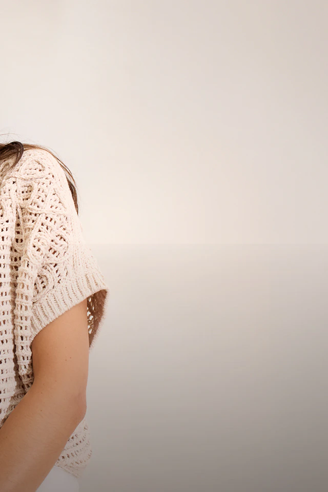 Frau trägt einen beigen gehäkelten Kurzarm Pullover mit einer weißen Hose.