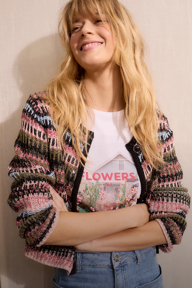 Frau trägt eine gemusterte Strickjacke aus rosa, gruen,blau, braun, schwarz, darunter ein bedrucktes Shirt und eine JEans.