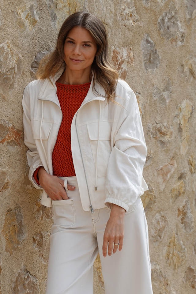 Raffaela Wachter trägt einen weissen Blouson mit einem gehäkelten orange-roten Pullover und einer 7/8 Hose in weiss.