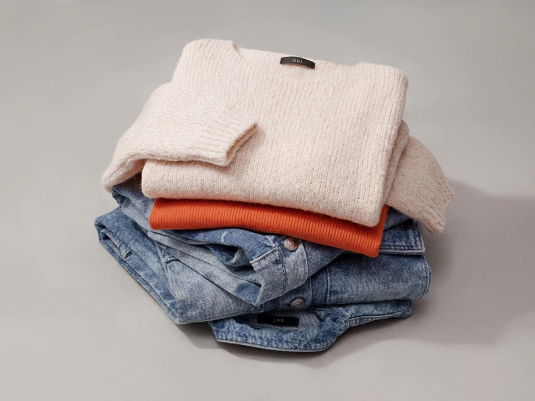 Stapel gefaltete Kleidung, oben ein beiger Strickpullover, dann ein orangenes Top, darunter Kleidung in Denim.