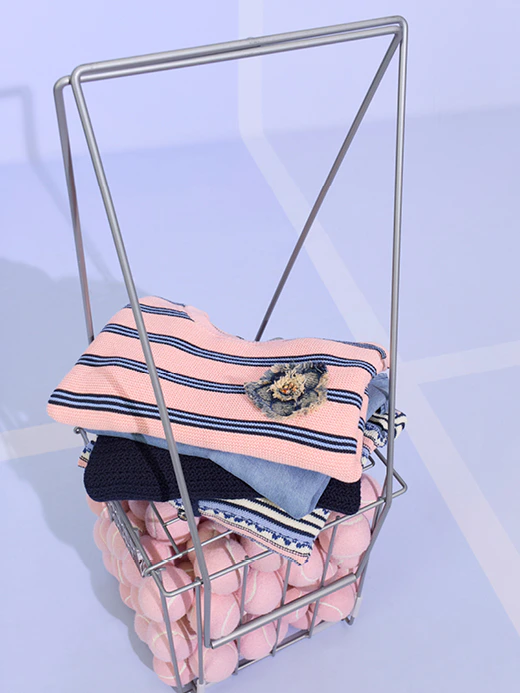 A basket containing pink tennis balls, with folded clothes on top. A pink jumper with blue stripes, underneath a light blue top, then a dark blue jumper.