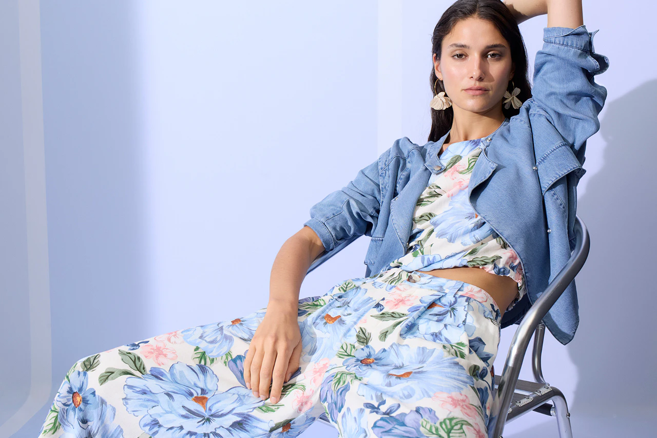 A woman is sitting, wearing an open denim jacket over a blouse with a floral print in pastel shades, and matching loose-fitting trousers in the same pattern.