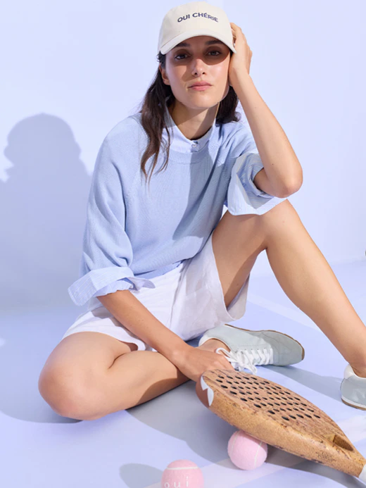 A woman sitting down wearing a light blue jumper; underneath, you can see a light blue shirt and white shorts.