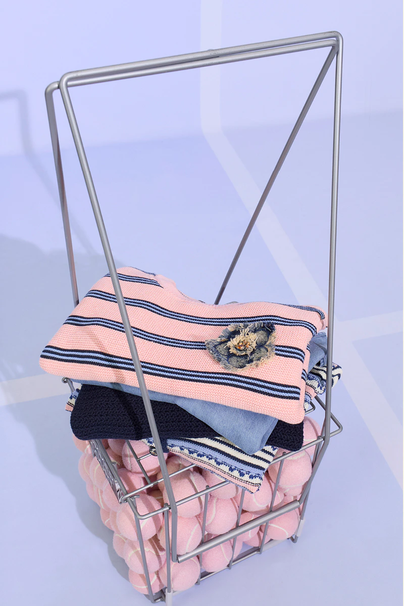 A basket containing pink tennis balls, with folded clothes on top. A pink jumper with blue stripes, underneath a light blue top, then a dark blue jumper.