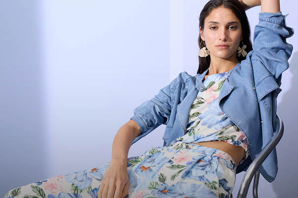 Frau sitzend trägt eine Jeansjacke offen, darunter eine Bluse mit einem blumendruck in pastelligen tönen und die passende lockere Hose in gleichem Muster.