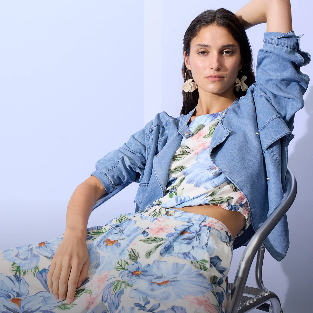 Frau sitzend trägt eine Jeansjacke offen, darunter eine Bluse mit einem blumendruck in pastelligen tönen und die passende lockere Hose in gleichem Muster.