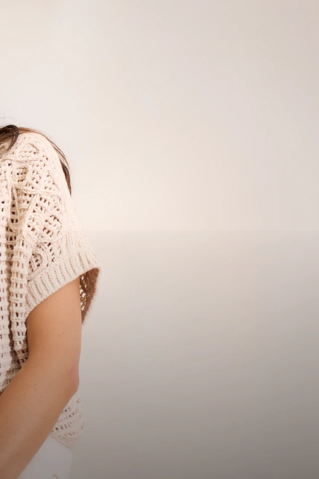 Frau mit einem beigen Pullover im Häkelstil und einer weißen Hose