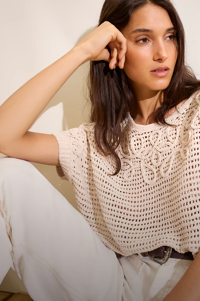 A woman wearing a beige crochet-style jumper and white trousers