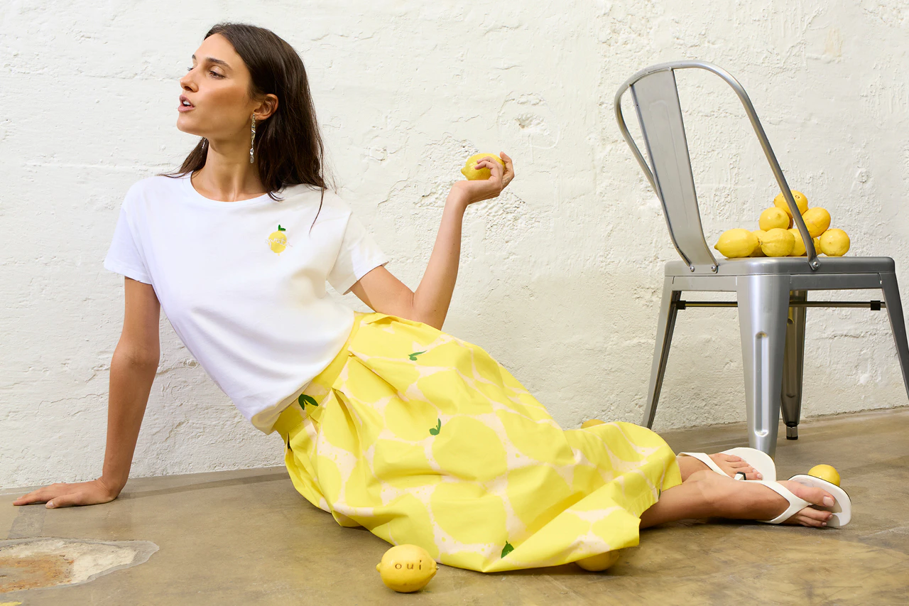 A woman wearing a white T-shirt with a lemon embroidered on the right breast, paired with a yellow skirt featuring a lemon print