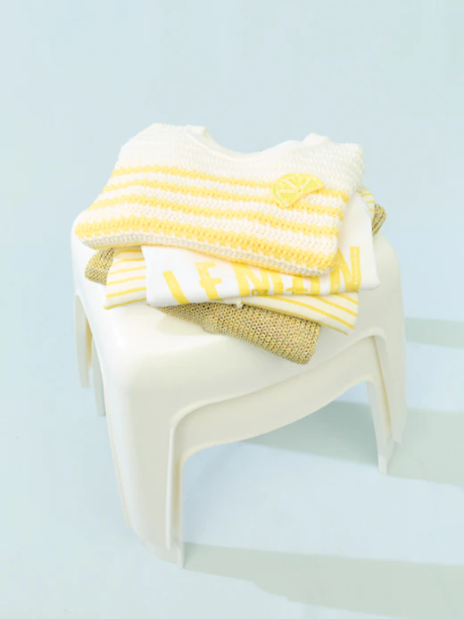 A white chair with folded clothes on it. You can see a yellow-and-white striped jumper, underneath which is a white T-shirt with the word ‘Lemon’ in yellow lettering, and other items of clothing in white and yellow.
