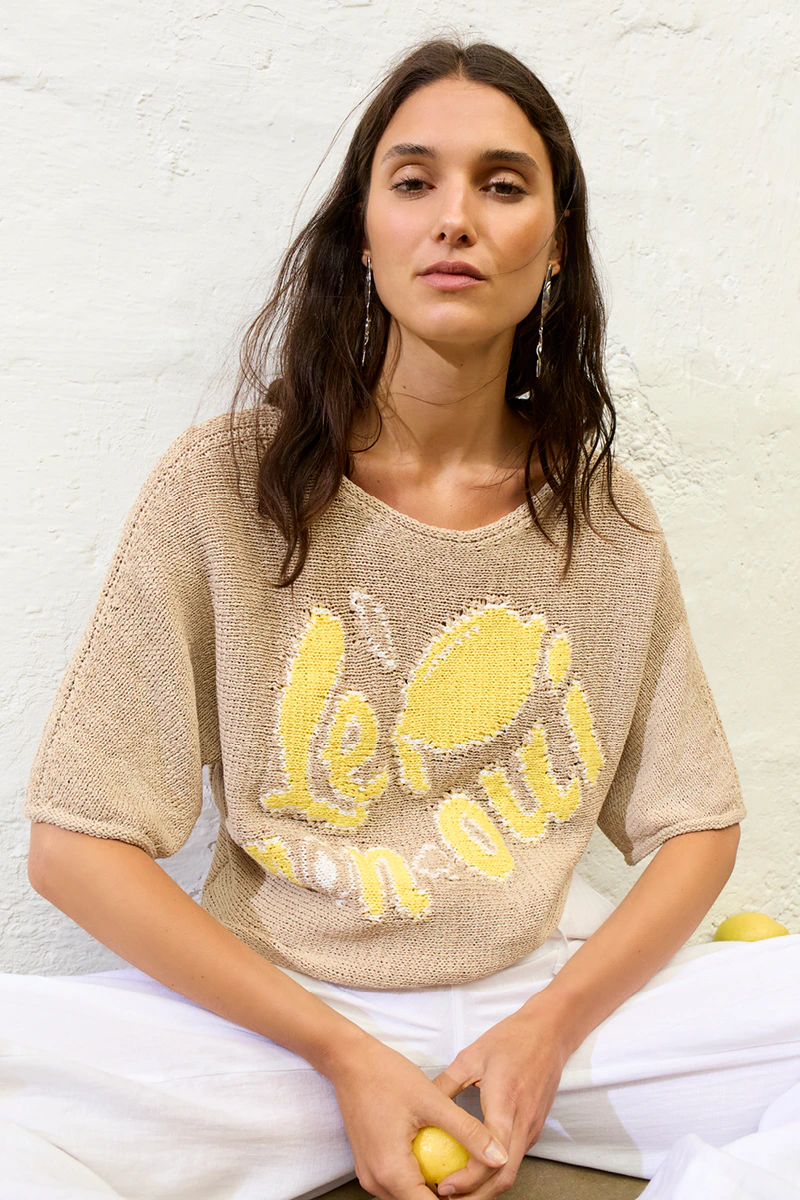 A woman is wearing a beige knitted jumper with a yellow lemon motif and lettering woven into the fabric, paired with white trousers.