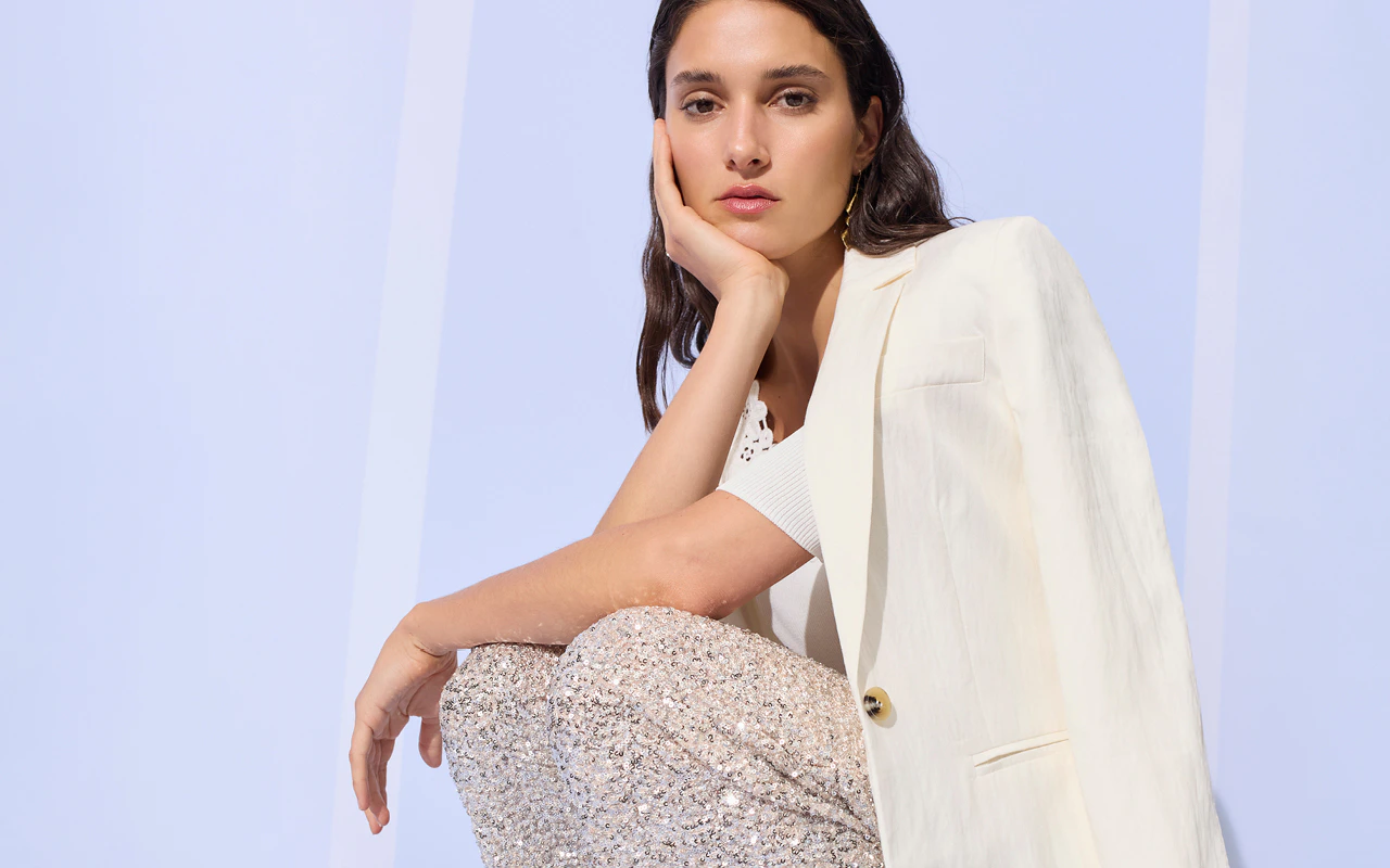 The woman is wearing a cream-coloured blazer, a white top and sequinned trousers.