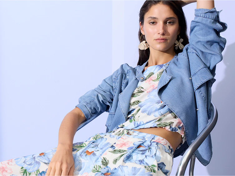 A woman is sitting, wearing an open denim jacket over a blouse with a floral print in pastel shades, and matching loose-fitting trousers in the same pattern.