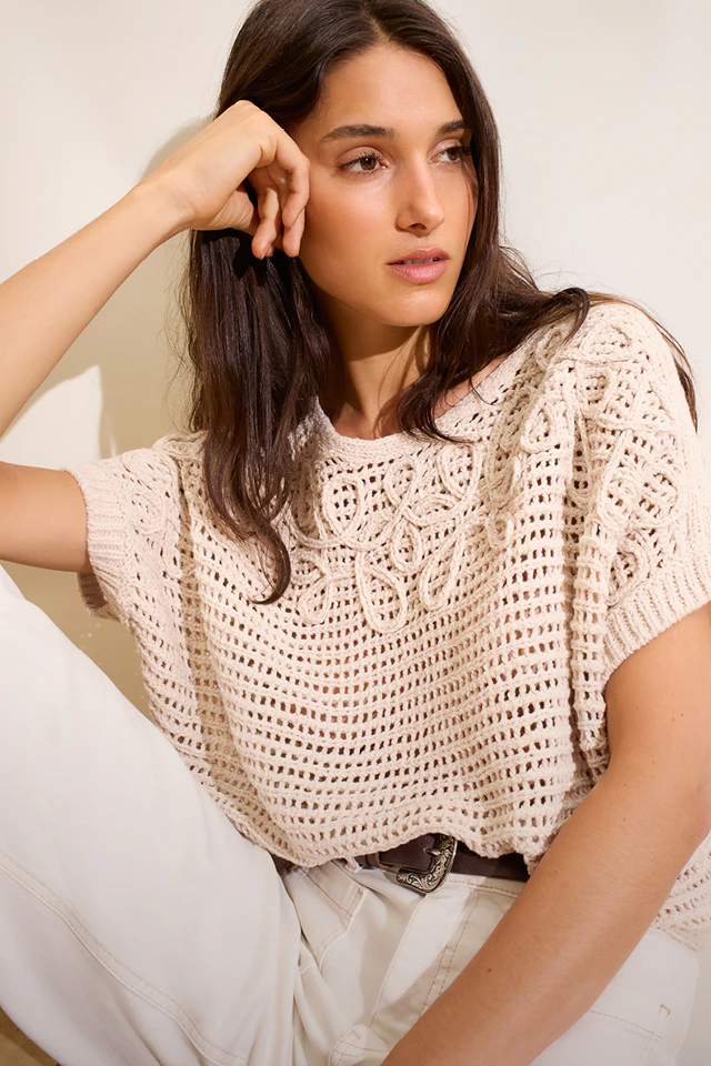 A woman is wearing a beige crochet top with white jeans.