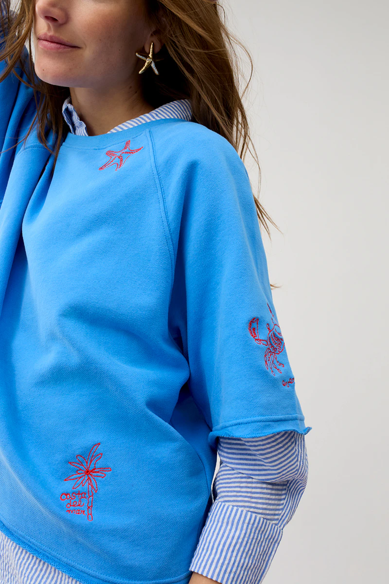 Frau trägt blauen Pullover mit aufgestickten roten Elementen aus dem Meer z.b. Krebs, Seestern und darunter eine blau-weiss gestreifte Bluse.