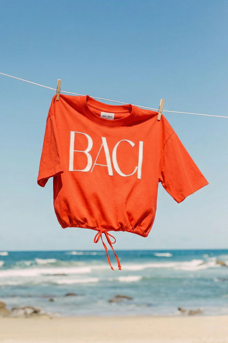 A red T-shirt hanging on a washing line by the sea, with the word ‘BACI’ written in white.
