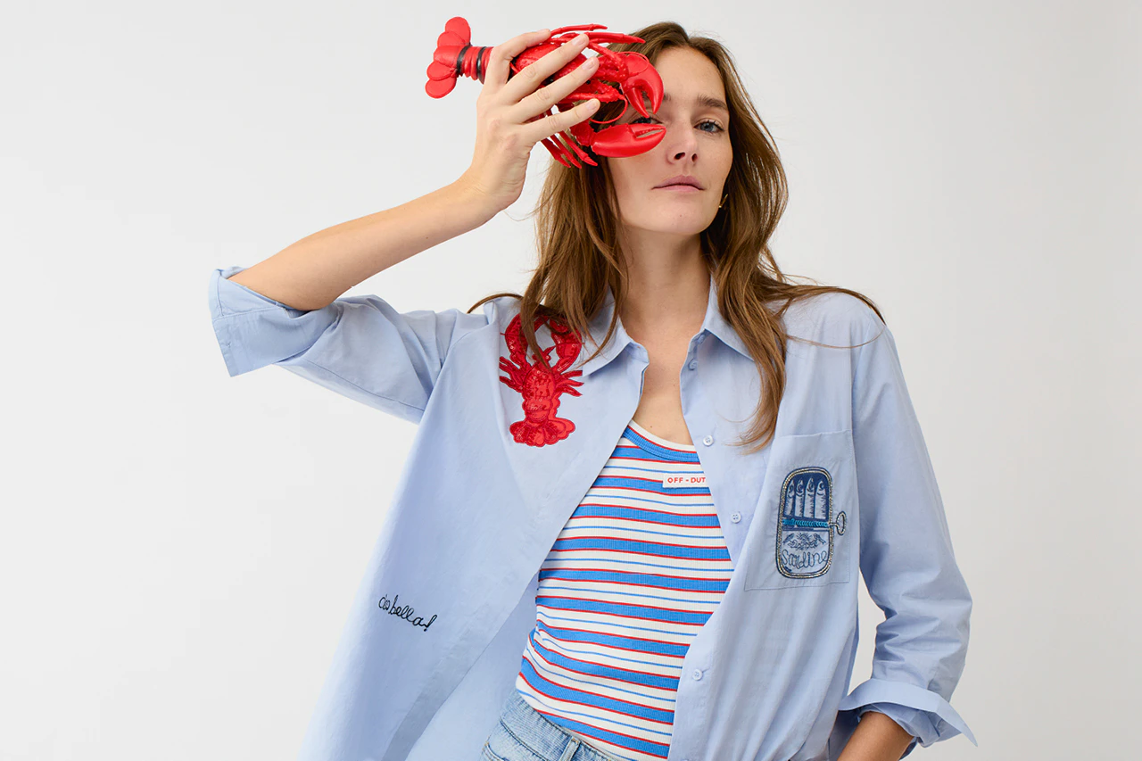 Frau trägt eine blaue Bluse mit Stickereien wie einen Krebs, oder eine Muschel, darunter ein Shirt in Streifen aus weiss, blau und rot und eine Jeans.