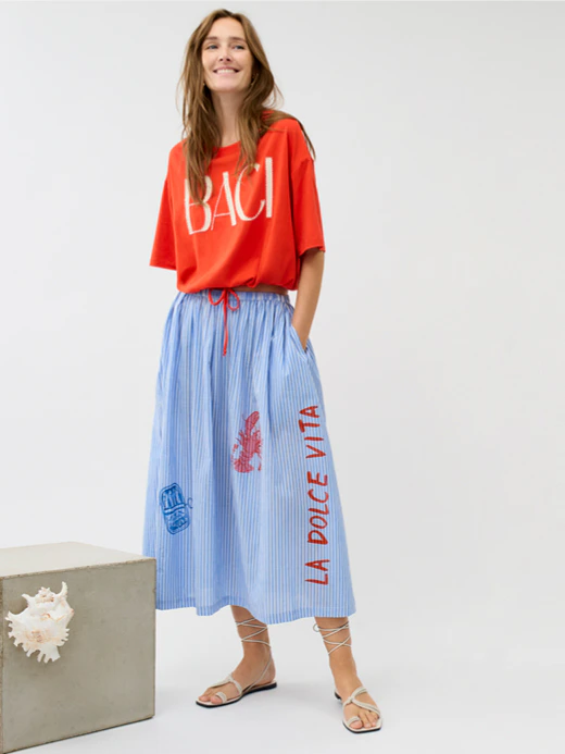 A woman is wearing a red T-shirt with the word ‘BACI’ in white lettering and a blue and white striped skirt featuring nautical prints such as crabs, mussels and lettering.