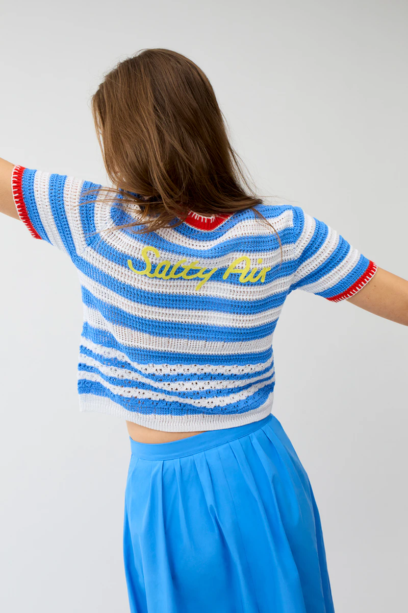 The woman, seen from behind, is wearing a blue and white striped short-sleeved jumper with red cuffs and yellow lettering on the back, paired with a blue skirt.