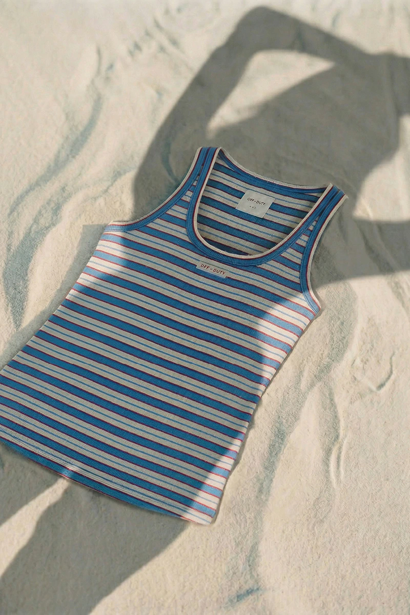A blue, white and red striped top lies on a patch of sand, and the silhouette of a woman wearing the top can be seen cast in the shade.