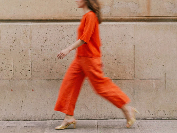 A woman in orange trousers and an orange blouse is walking from right to left. The image is slightly blurred due to movement.