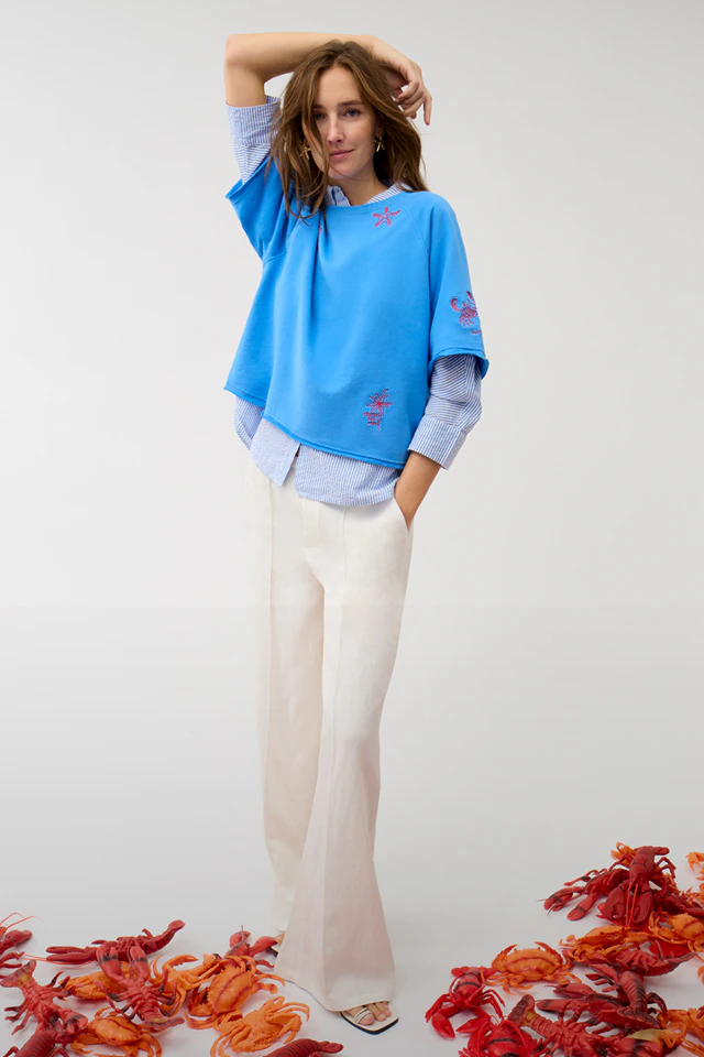 Frau trägt blauen Pullover mit aufgestickten roten Elementen aus dem Meer z.b. Krebs, Seestern und darunter eine blau-weiss gestreifte Bluse und einer weißen Jeans.