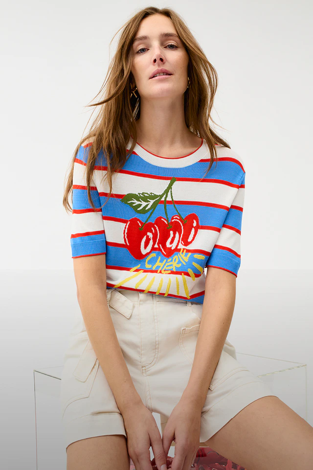 A woman is wearing a short-sleeved striped jumper in white, blue and red, featuring a knitted-in cherry and lettering, and white shorts.