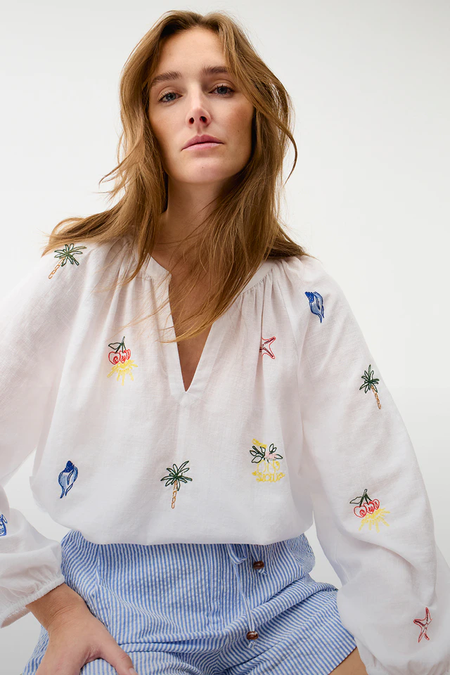 A woman is wearing a white blouse with colourful sea-themed embroidery and blue shorts.