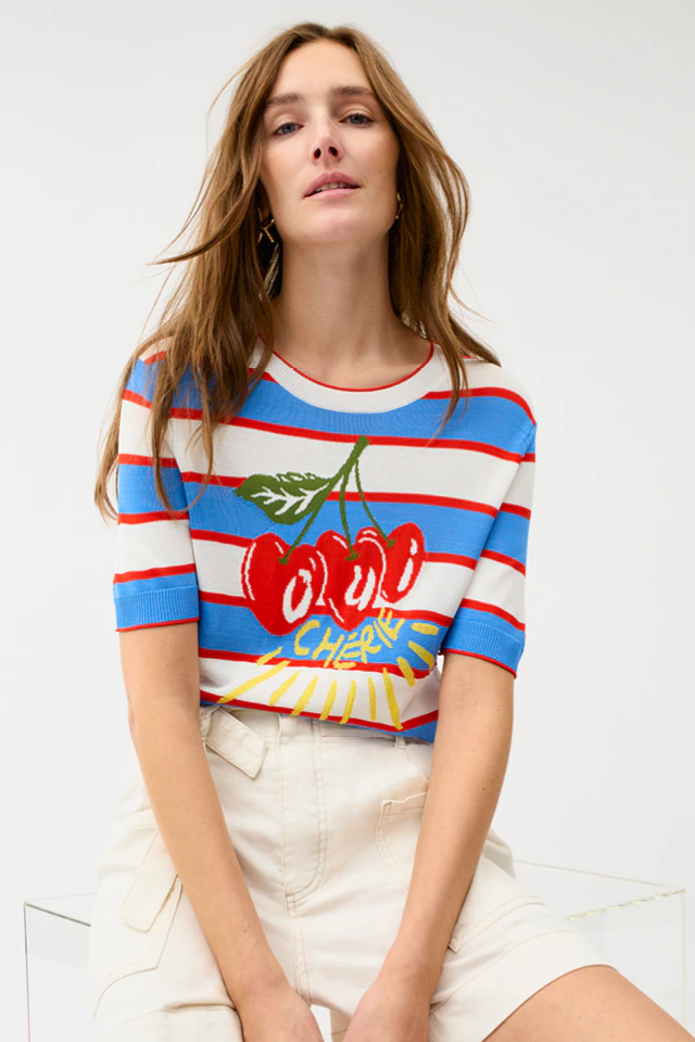 The model is wearing a blue, red and white striped T-shirt with cherries on it and cream-coloured Bermuda shorts. 