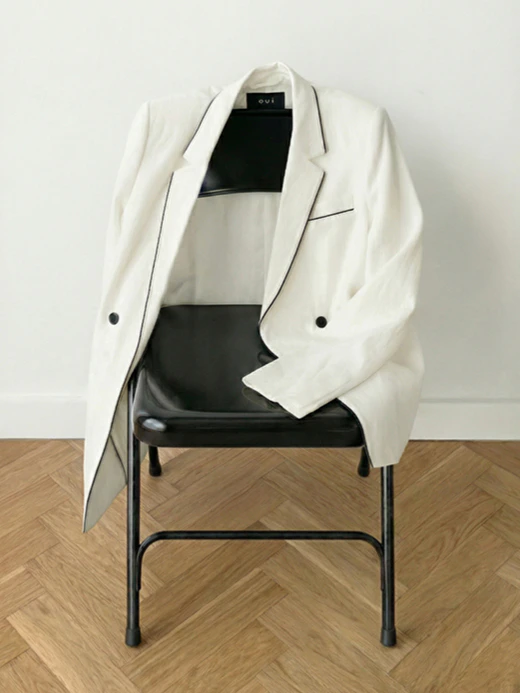 Ueber einer Stuhllehne hängt ein weisser Blazer mit schwarzem Piping.