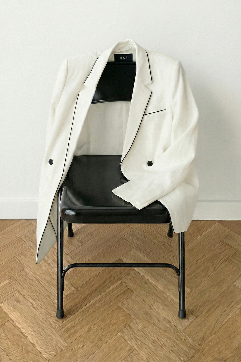 Ueber einer Stuhllehne hängt ein weisser Blazer mit schwarzem Piping.