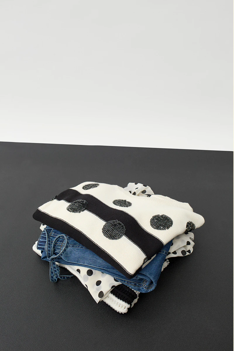 Ein Stapel gefalteter Kleidung, oben eine weiss schwarz gestreifter Pullover mit schwarzen Punkten, darunter ein Jeansteil und darunter ein Shirt in weiss mit schwarzen Punkten.