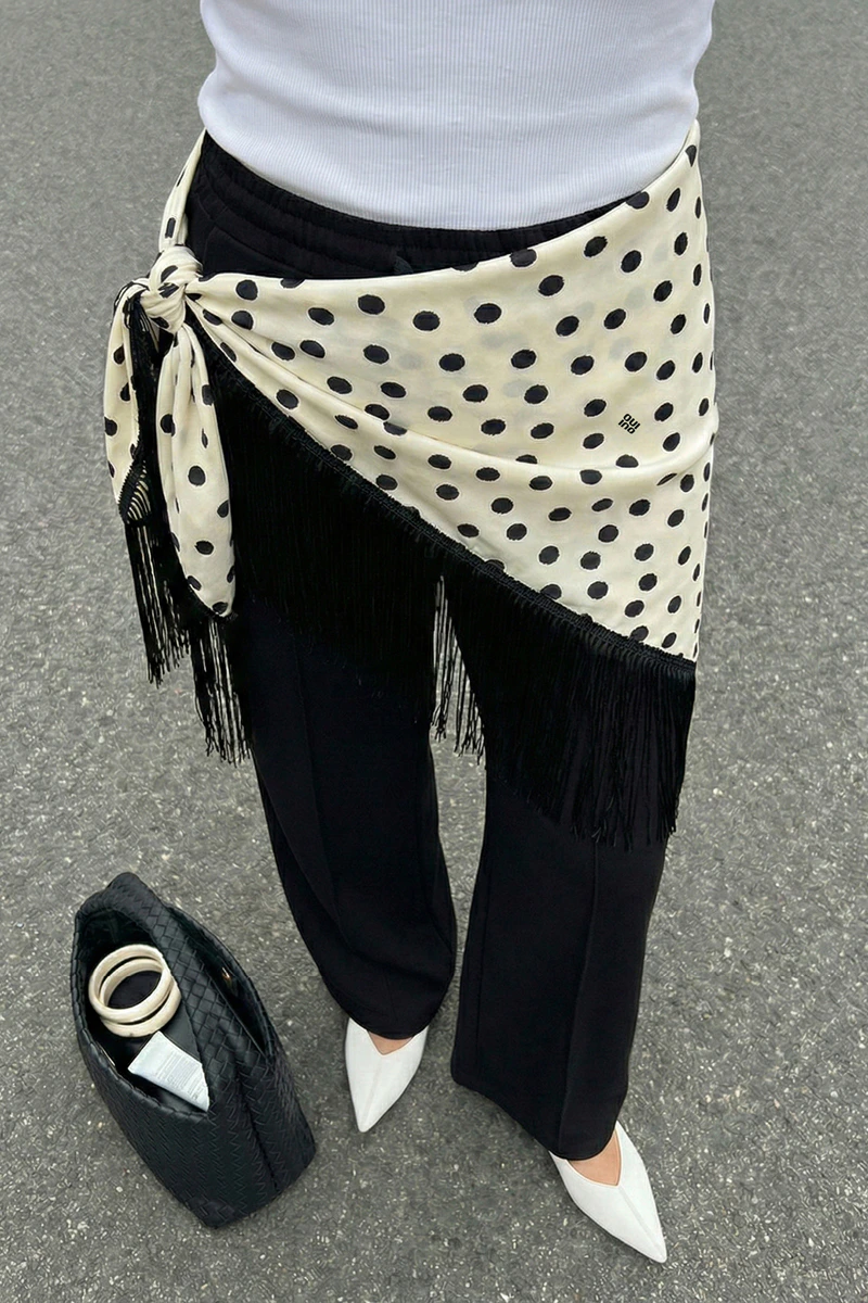 Eine Aufnahme ab Bauch abwärts einer Frau. Sie trägt ein weisses Top, eine schwarze Hose und über der Hose ein dreieckstuch in weiss mit schwarzen Punkten und schwarzen FRansen.