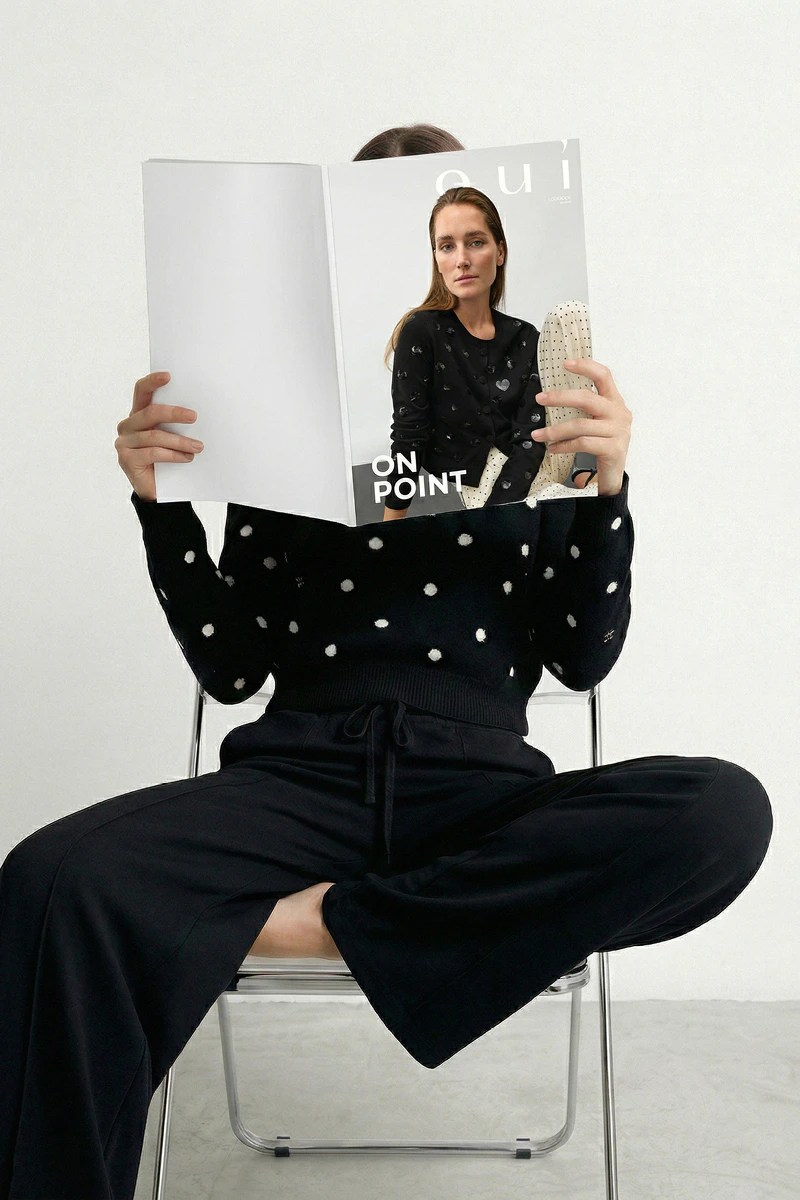 A woman is sitting on a chair with a magazine in front of her face; you can see a black polka-dot jumper and black trousers.