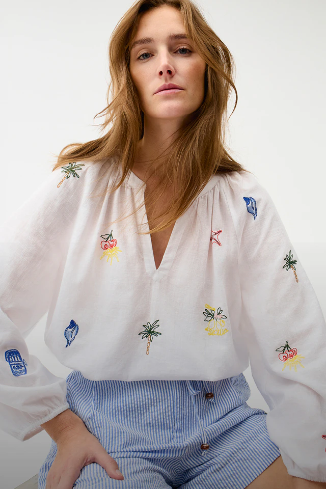 A woman is wearing a white blouse with colourful sea-themed embroidery and blue shorts.