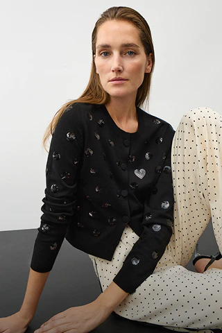 A woman is wearing a black cardigan with black sequinned dots, paired with white trousers with black dots.