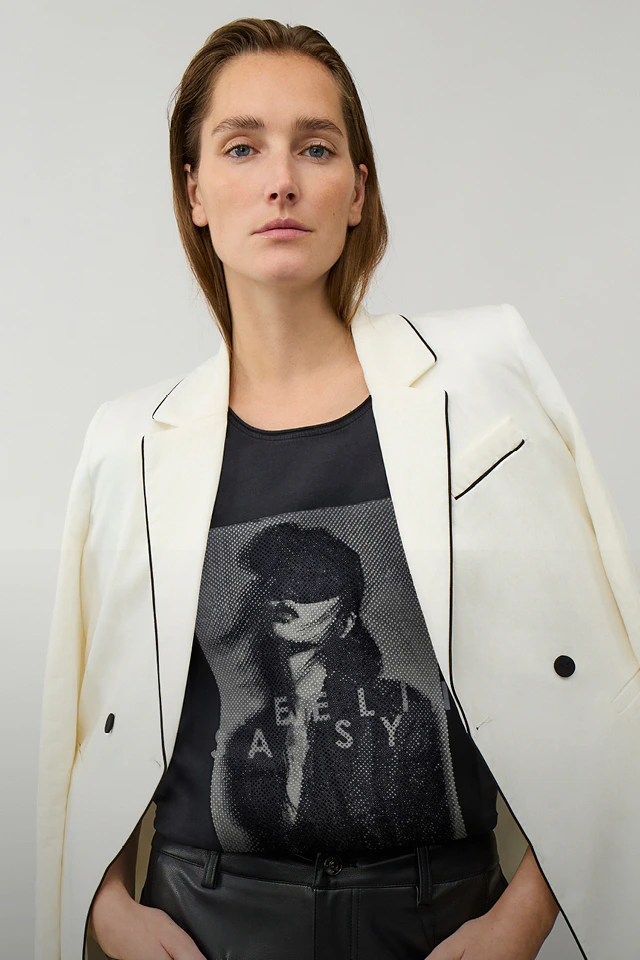 Frau trägt einen weißen Blazer mit schwarzen Kontrastnähten um die Schultern gehängt, darunter ein schwarzes Shirt mit einem grauen Aufdruck, dazu einen passende schwarze Hose.