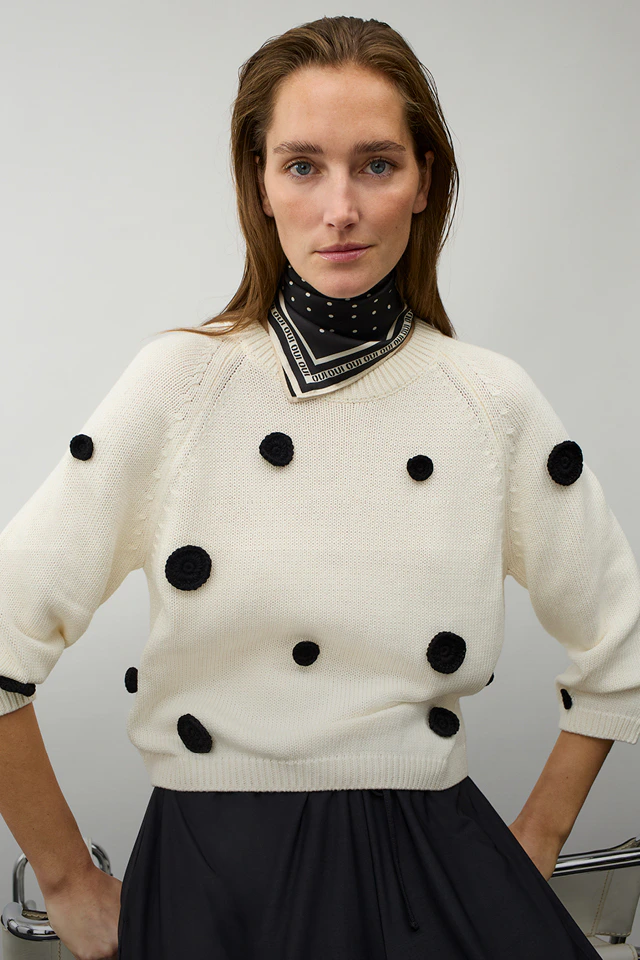 Frau mit weißem Pullover mit schwarzen Punkten, schwarz-weissem Halstuch und schwarzem Rock.