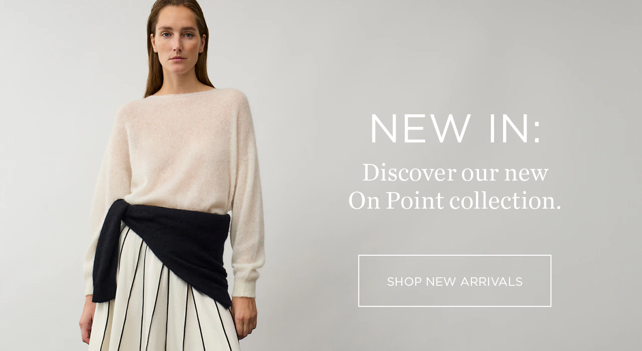 The model is wearing a white jumper and a white pleated skirt with black piping, and has a black jumper tied around her waist.