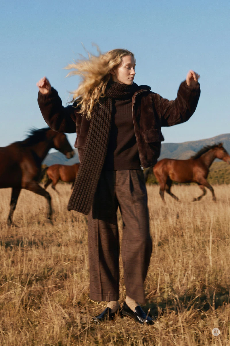Herbstliche Damenmode: Model trägt eine braune Jacke, einen dunklen Strickpullover mit langem Schal, eine braune karierte Hose und schwarze Loafer. Sie steht auf einem Feld, während im Hintergrund mehrere Pferde laufen und Berge unter blauem Himmel zu sehen sind.