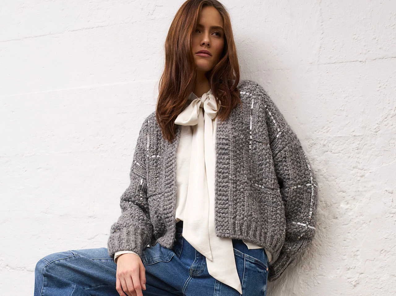 Model trägt eine graue Strickjacke mit Pailletten, eine cremefarbene Bluse mit Schleifenkragen und eine blaue Jeans aus der Oui Heritage Reset Kollektion.