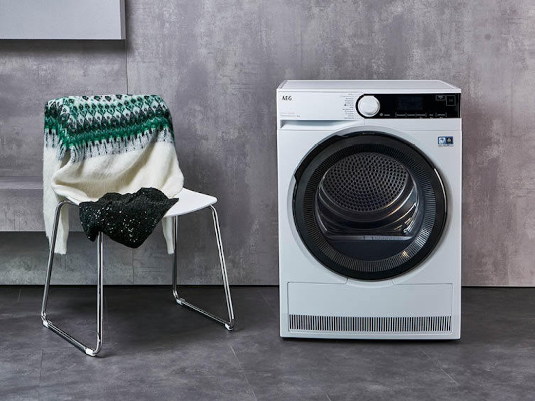 A white washing machine stands in front of a grey wall. Next to it is a white chair with a green and white knitted jumper and a dark item of clothing draped over the back.