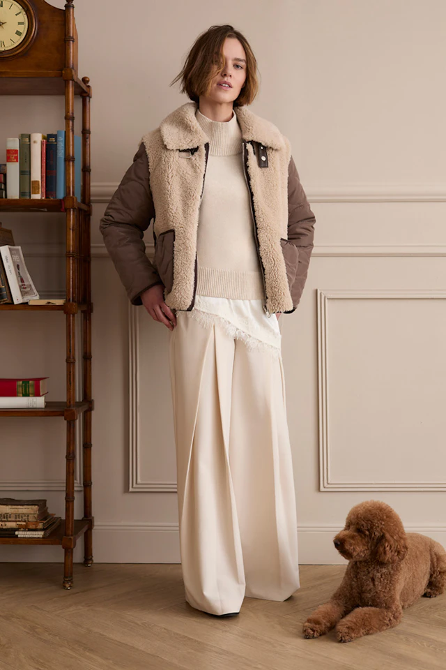 Model trägt eine braune Jacke mit Teddyfutter über einem cremefarbenen Strickpullover und einer weiten cremefarbenen Hose. Neben ihr liegt ein brauner Hund.