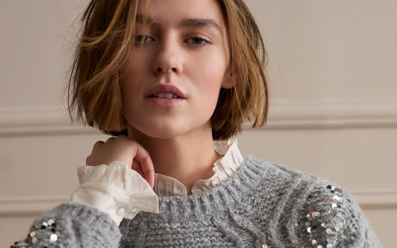 A light grey knitted jumper with sequinned detailing on the shoulders, paired with a white blouse featuring a feminine ruffled collar.