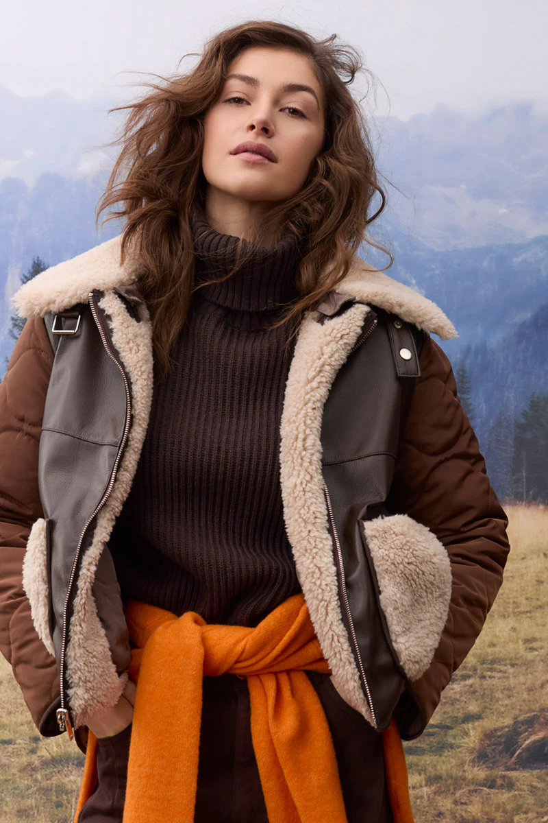 Damenmode im Herbst: Model trägt eine braune Jacke mit hellem Teddyfell-Besatz über einem dunkelbraunen Strickpullover. Ein orangefarbener Pullover ist um die Taille gebunden. Im Hintergrund ist eine alpine Berglandschaft mit Wald und Wiesen zu sehen.