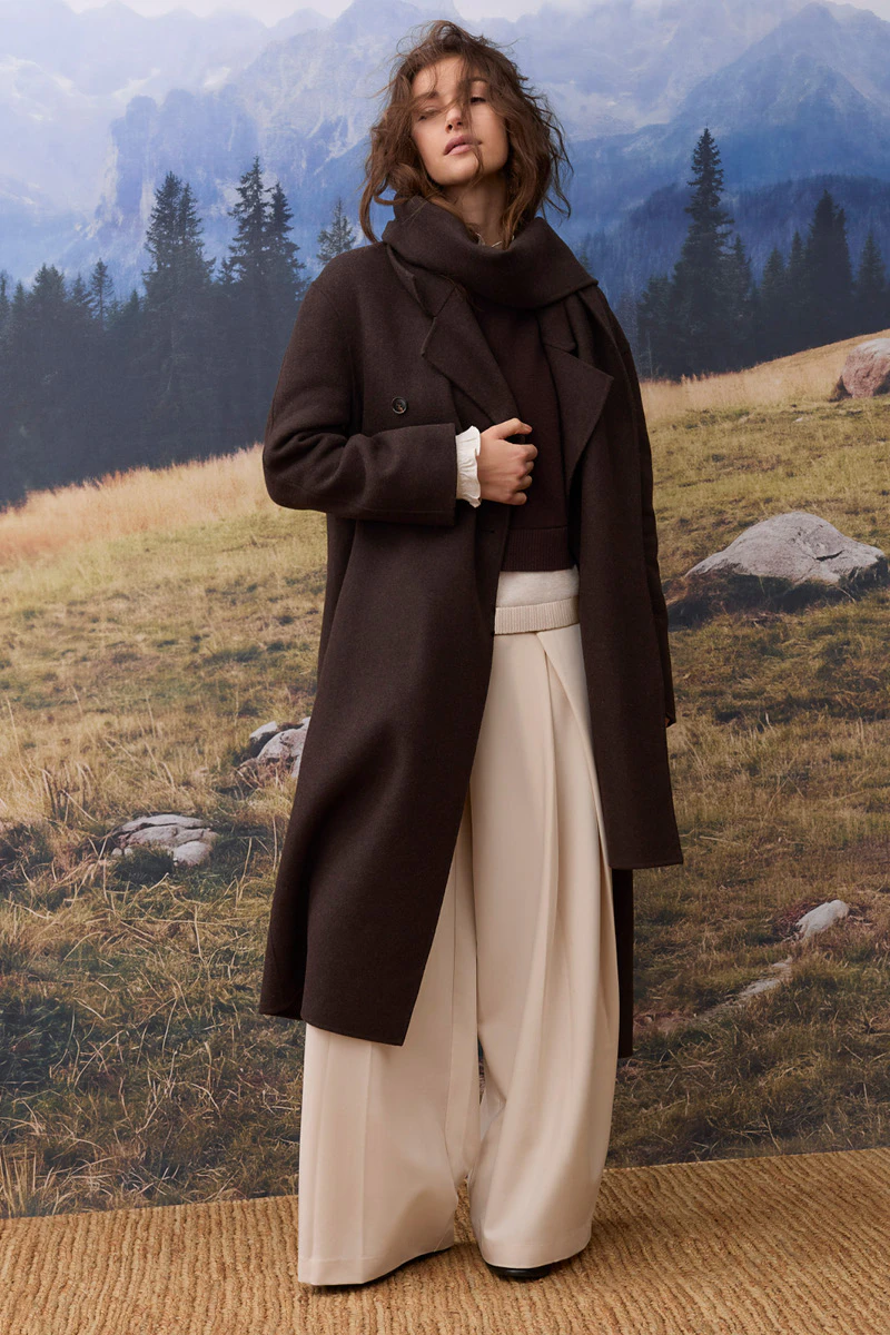 Damenmode im Herbst: Model trägt einen langen dunkelbraunen Mantel mit passendem Schal über einem braunen Pullover und einer weit geschnittenen cremefarbenen Hose. Im Hintergrund ist eine alpine Berglandschaft mit Wald, Wiesen und Felsen zu sehen.