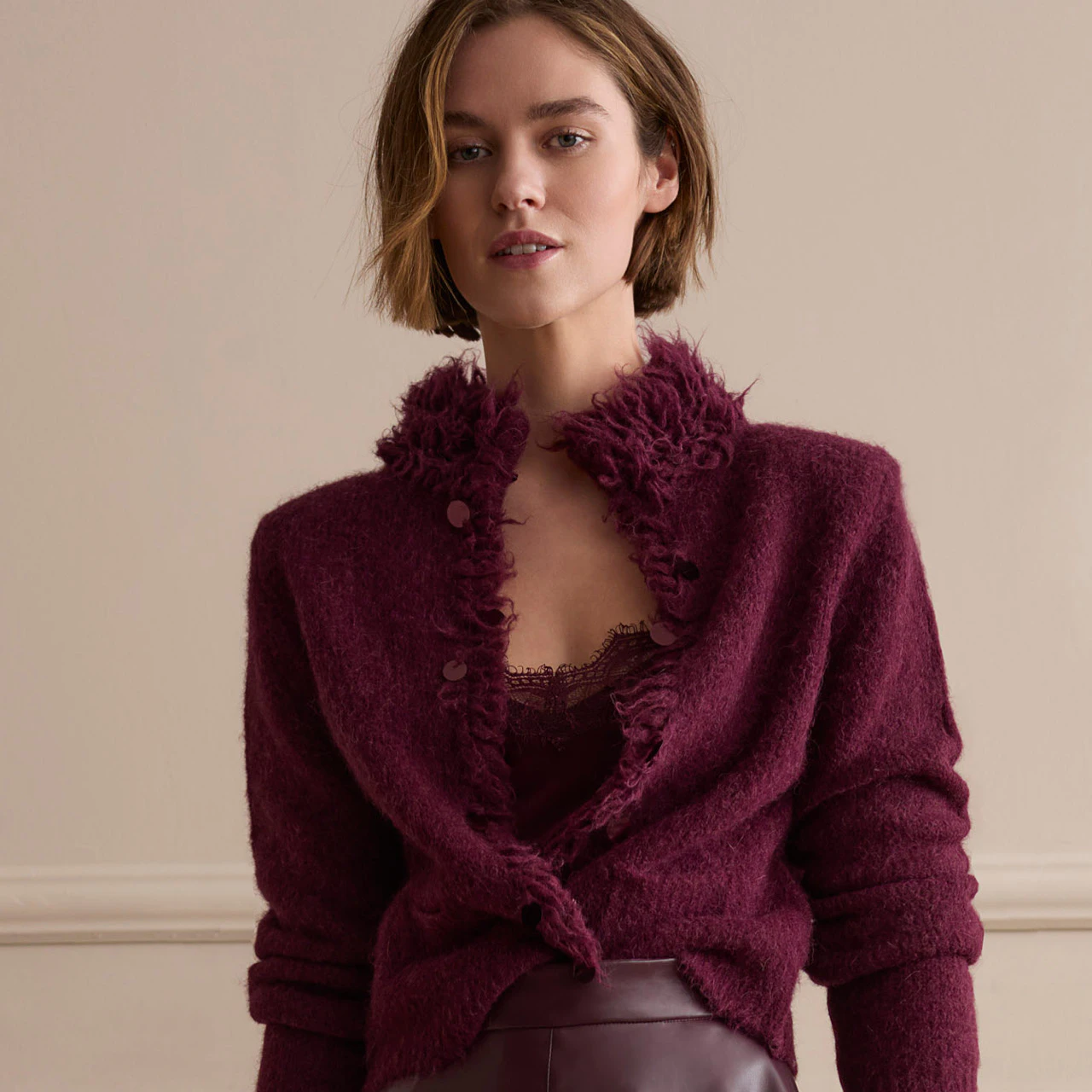 The model is wearing a burgundy cardigan with fringed detailing and a matching top, paired with a dark red leather midi skirt.